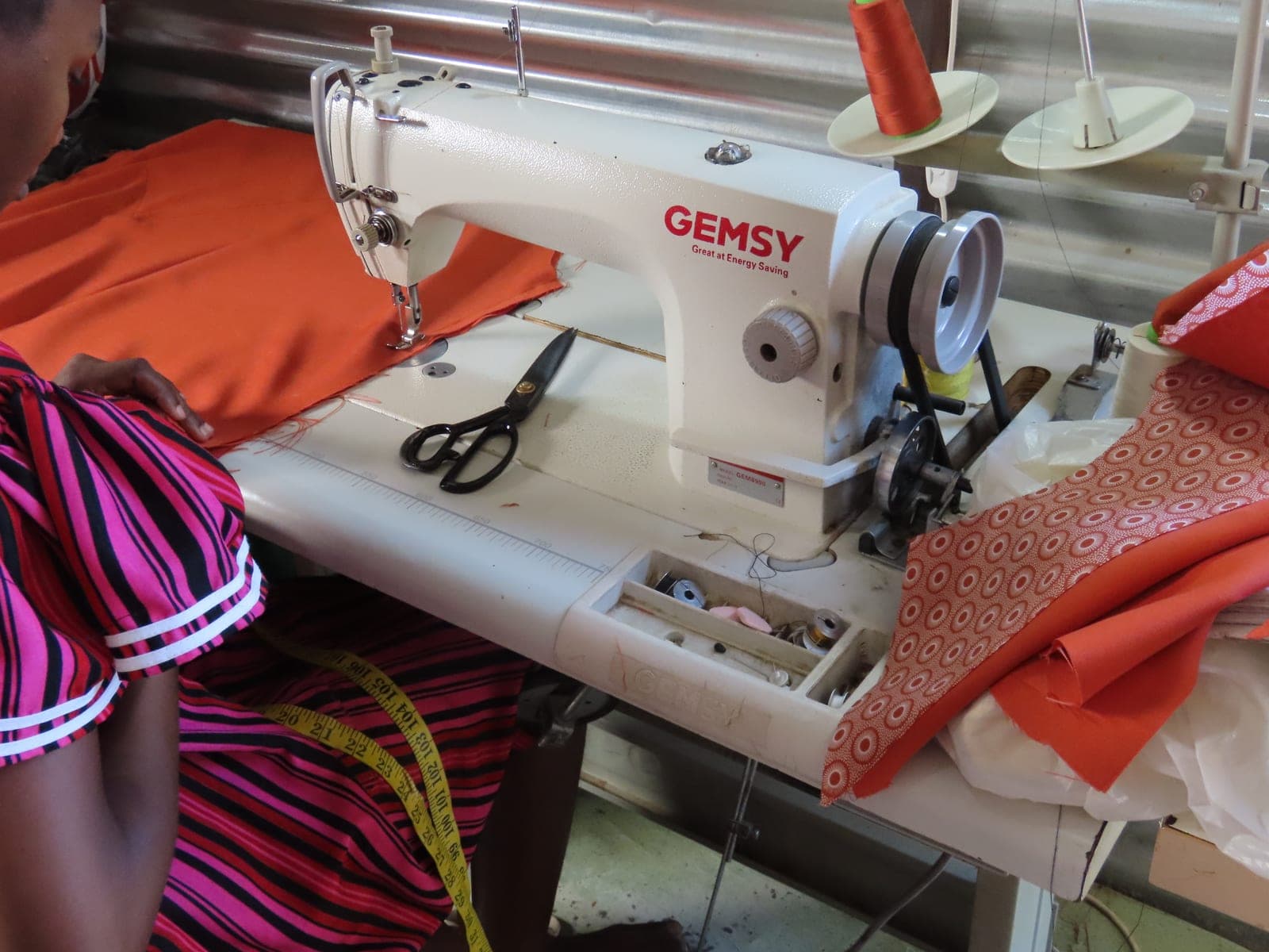Tailor stitching orange fabric at an industrial sewing machine with scissors and measuring tape in a boutique workshop