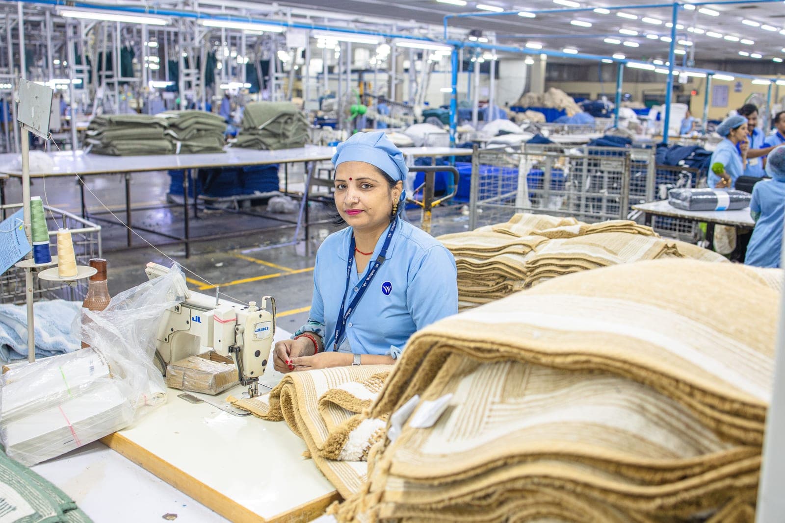Garment factory worker at an industrial sewing machine with stacks of cut pieces on a busy production floor