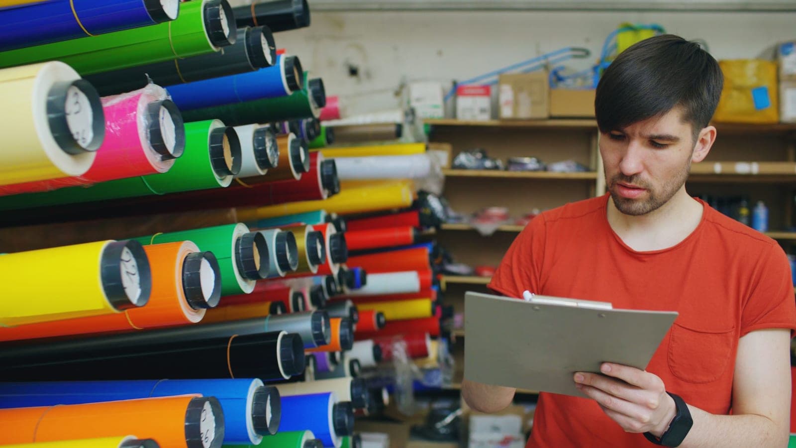 A person checking a clipboard against rows of material rolls while managing stock and orders