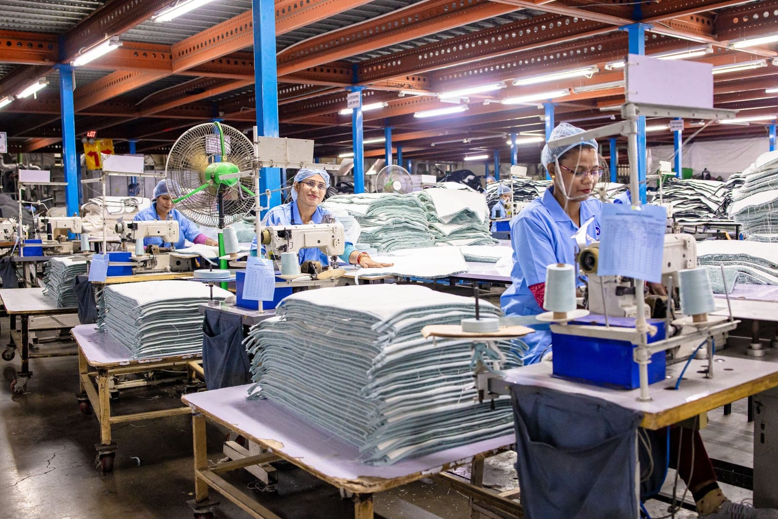 Textile production floor with workers at sewing machines and stacks of cut fabric, showing production in progress