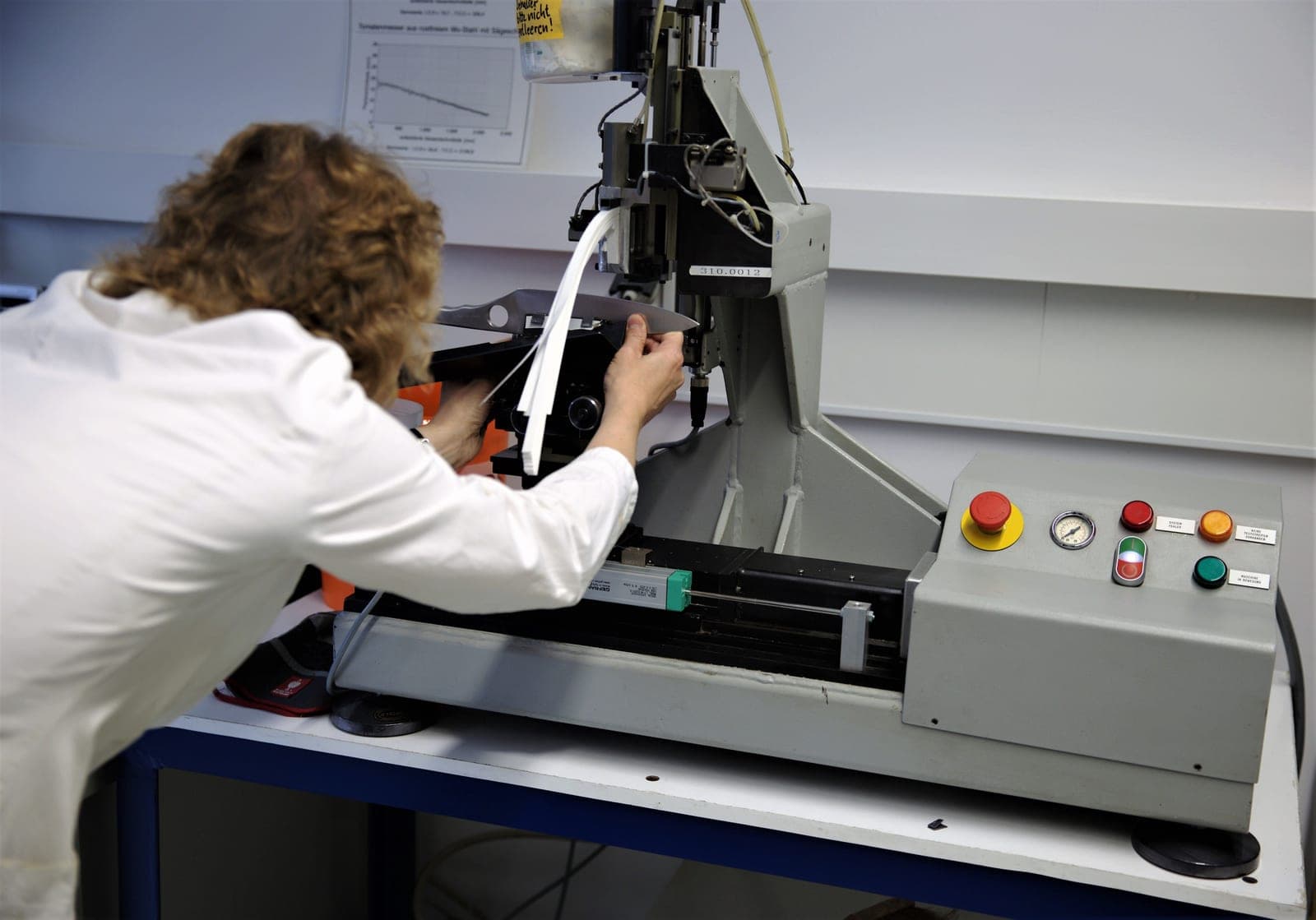 A technician in a lab coat operating a materials testing machine with a strength-test graph on the wall